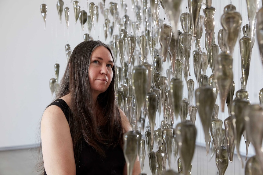 The artist Yhonnie Scarce standing next to her sculpture made of a thousand glass yams hanging from the ceiling