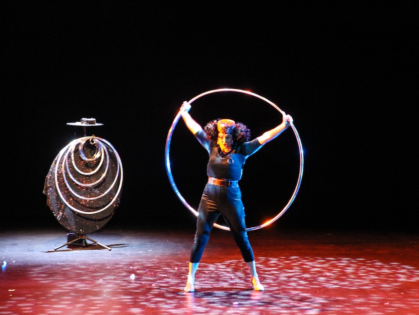 A woman in black holds a big silver hoop behind herself with several smaller hoops hanging up on the side of the stage.