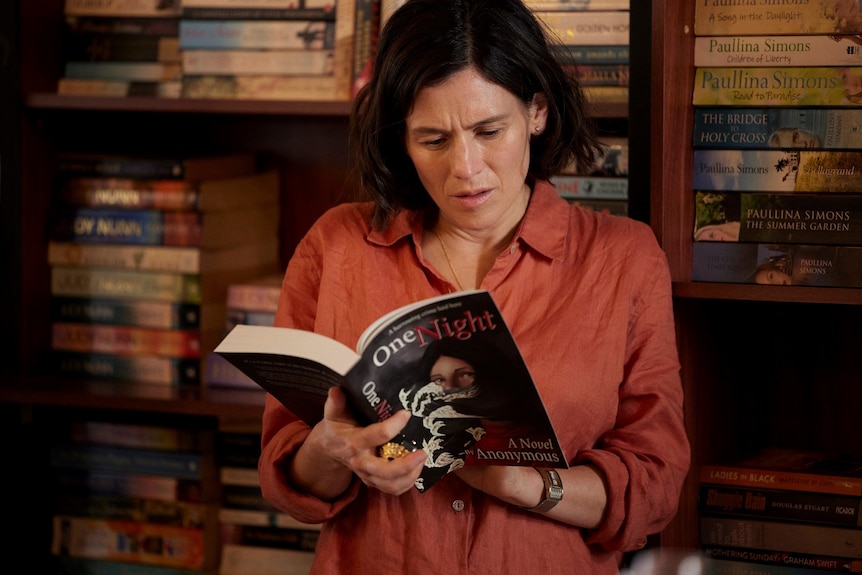 Yael Stone as Hat engrossed in an open book she has in her hands called One Night, written by Anonymous