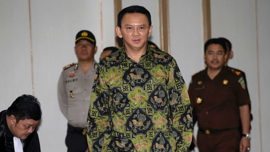 A shot of Ahok walking with officials in the background. Ahok wears glasses, a shirt and trousers.