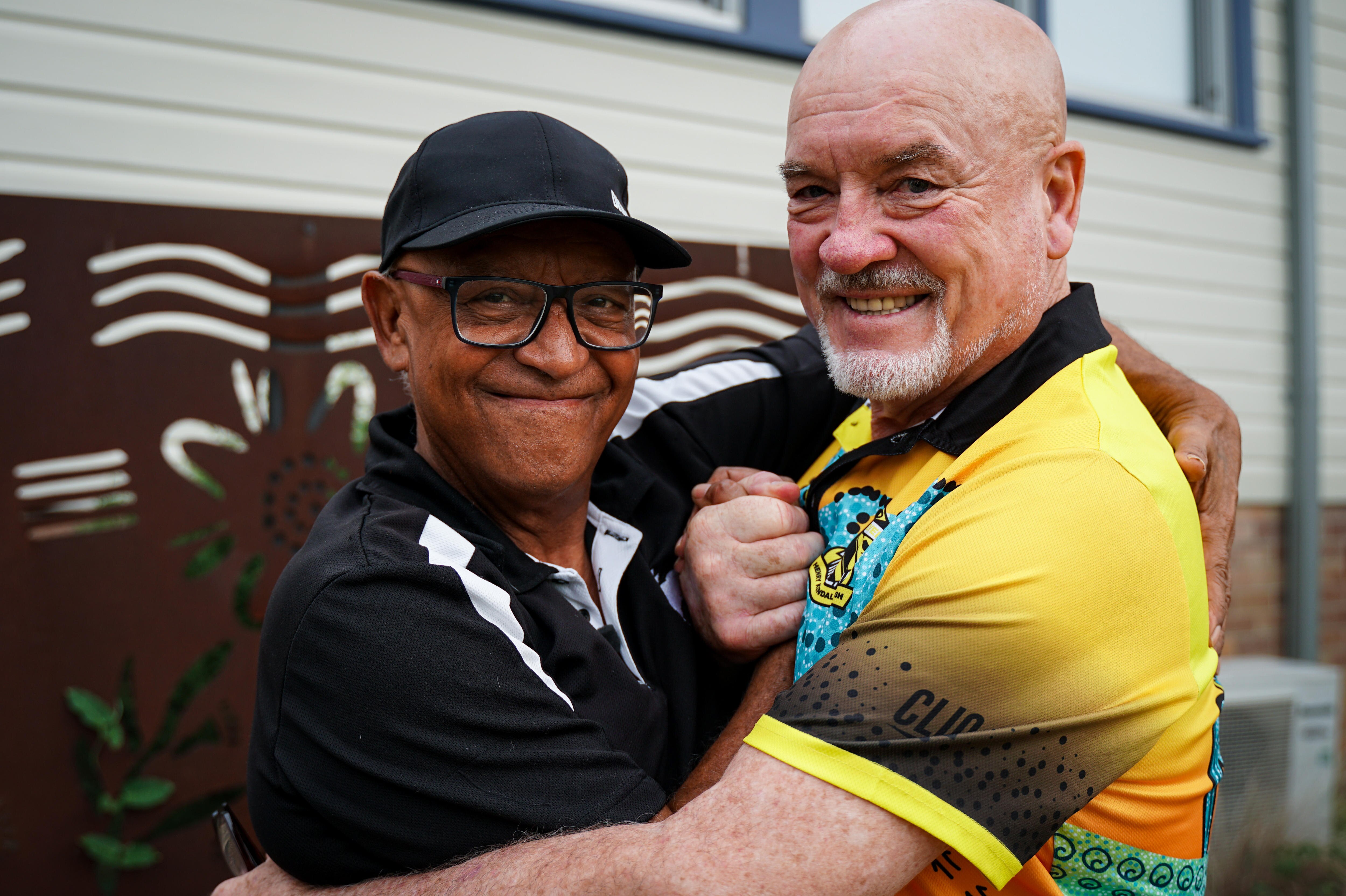Two men hugging and smiling at the camera, one wearing black cap, black and white tee.