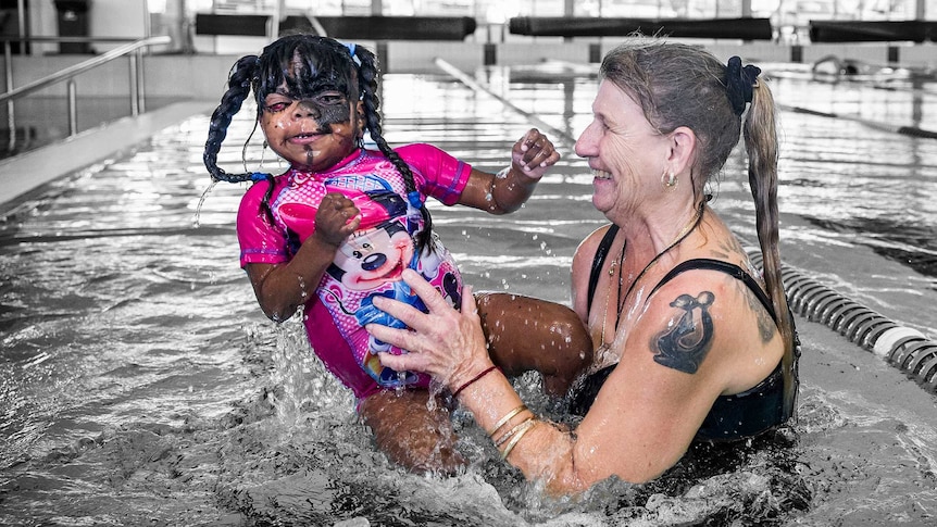 A girl in a Minnie Mouse swimsuit is lifted out of a pool by her mother. The people are in colour, background black and white