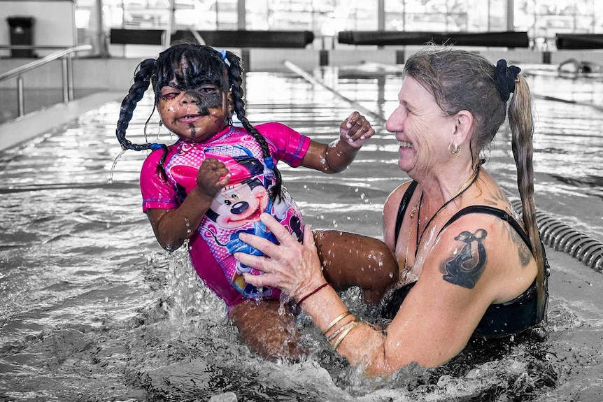 A girl in a Minnie Mouse swimsuit is lifted out of a pool by her mother. The people are in colour, background black and white