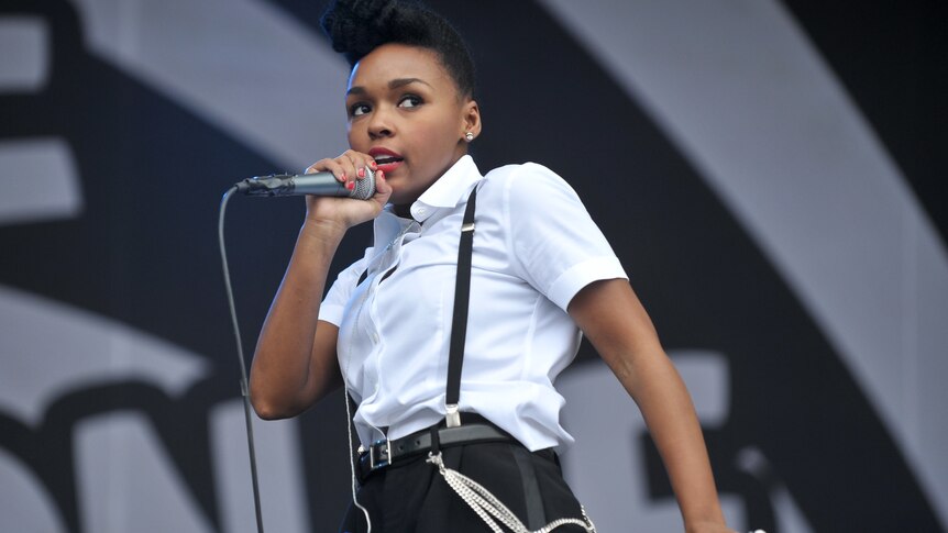 Janelle Monae wears a white shirt with black pants and suspenders. She sings into a microphone on stage.