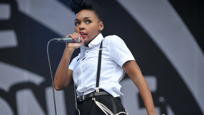 Janelle Monae wears a white shirt with black pants and suspenders. She sings into a microphone on stage.