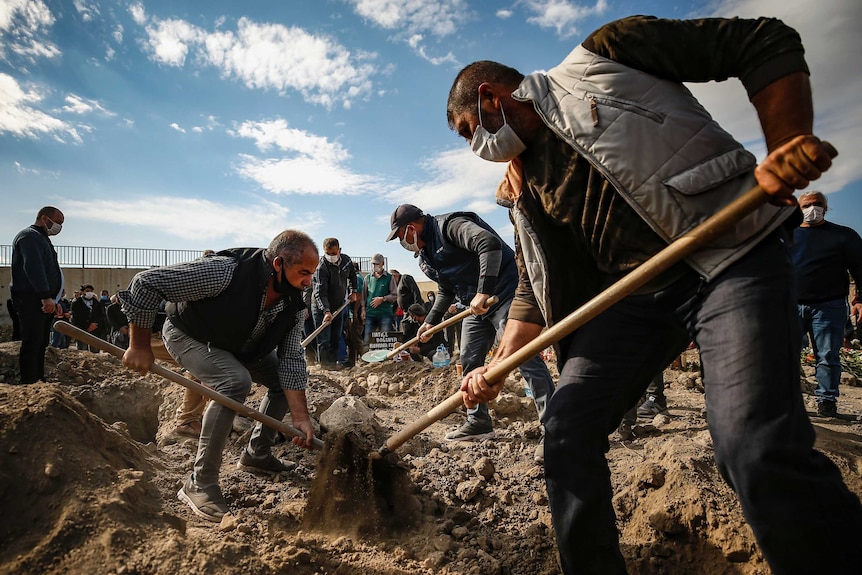 Men wearing face masks cover graves in soil under a blue sky.