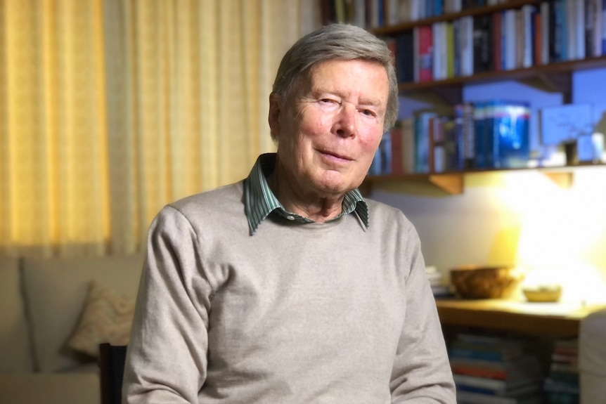 An old man in a grey sweater smiles at the camera in front of a shelf of books