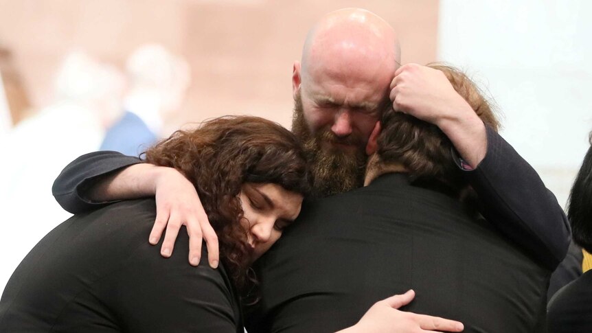 A close-up shot shows three people clad in black embracing, with a man at the centre crying.