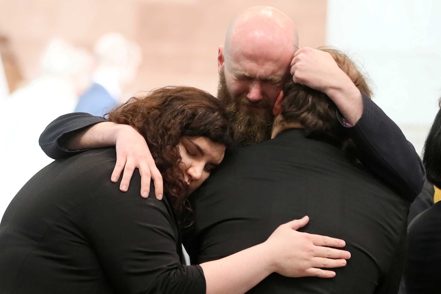 A close-up shot shows three people clad in black embracing, with a man at the centre crying.