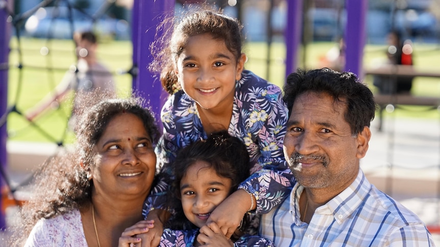 A Tamil family of four sits at a playground with big smiles - mum, dad and two little girls