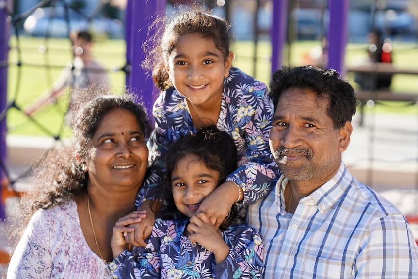 Тамильская семья из четырех человек сидит на детской площадке и широко улыбается - мама, папа и две маленькие девочки.