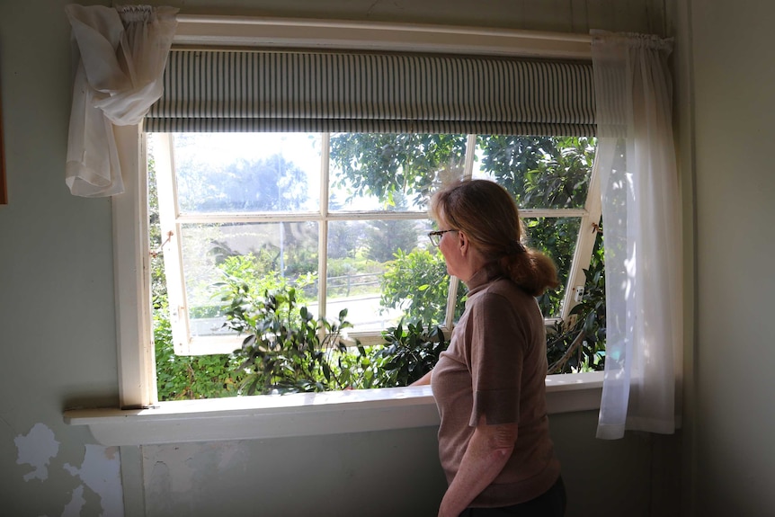 A woman opens the window of a cottage, looking out at trees in the garden