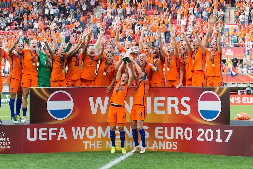 A women's soccer team wearing orange and blue lifts a trophy
