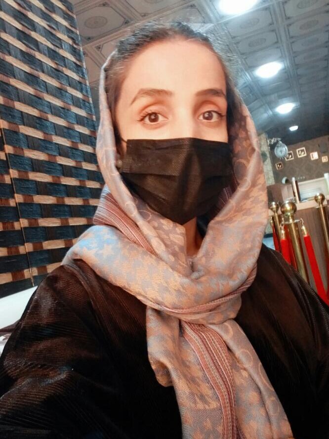 A woman wearing a head scarf and black face mask looks into camera as she takes a selfie