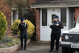 Police officers stand guard with a police vehicle next to them at the front of a house.