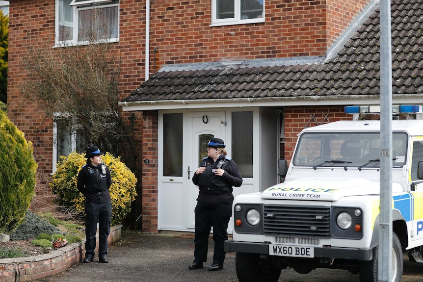 Police officers stand guard with a police vehicle next to them at the front of a house.