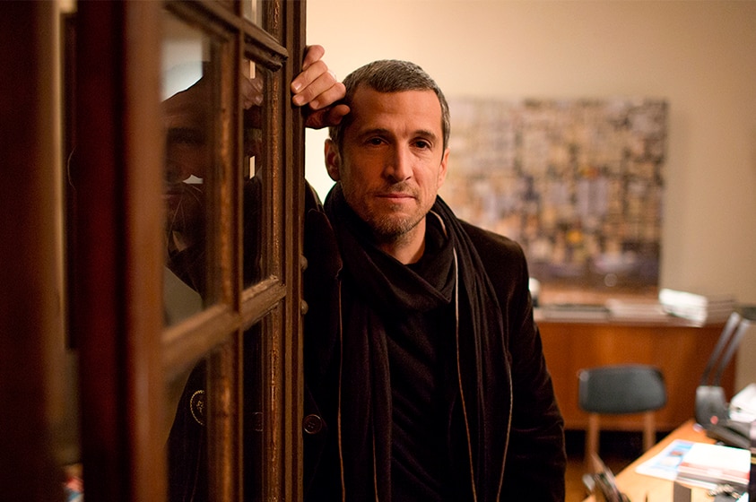 A man with grey buzzcut wears dark coat and scarf and stands and leans with raised hand on interior door in warmly lit room.