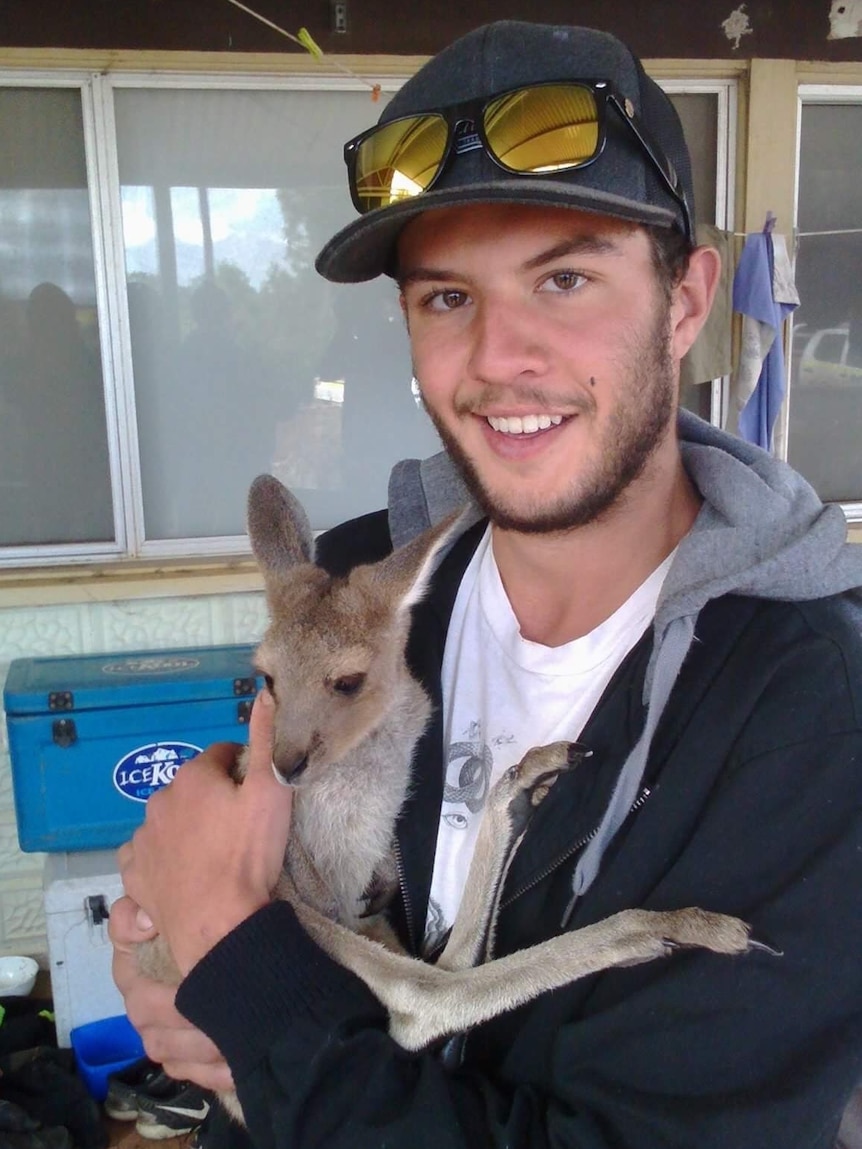 A young man with a light beard smiles while holding a small marsupial.
