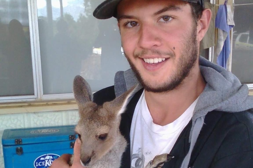 A young man with a light beard smiles while holding a small marsupial.