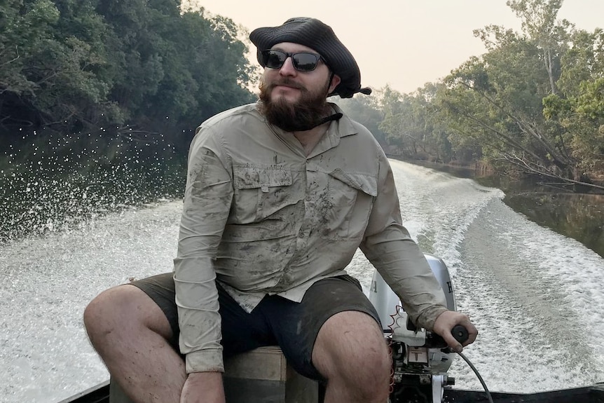 Bearded man, black cap, glasses, steers boat on remote creek, wears long sleeve shirt, black shorts. Slight smile.