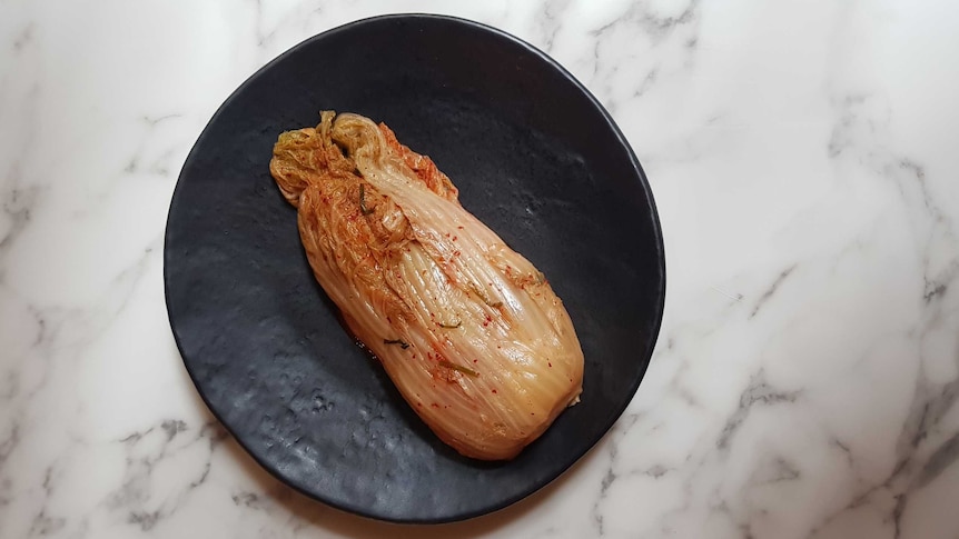 A whole wombok cabbage kimchi on a black ceramic plate, sitting on a marble benchtop.