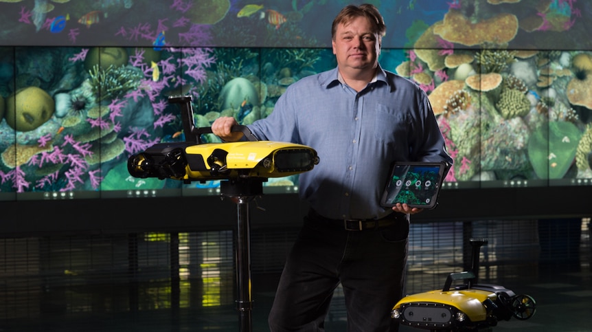 Matt Dunbabin stands next to two yellow robots in a room with a coral picture backdrop.