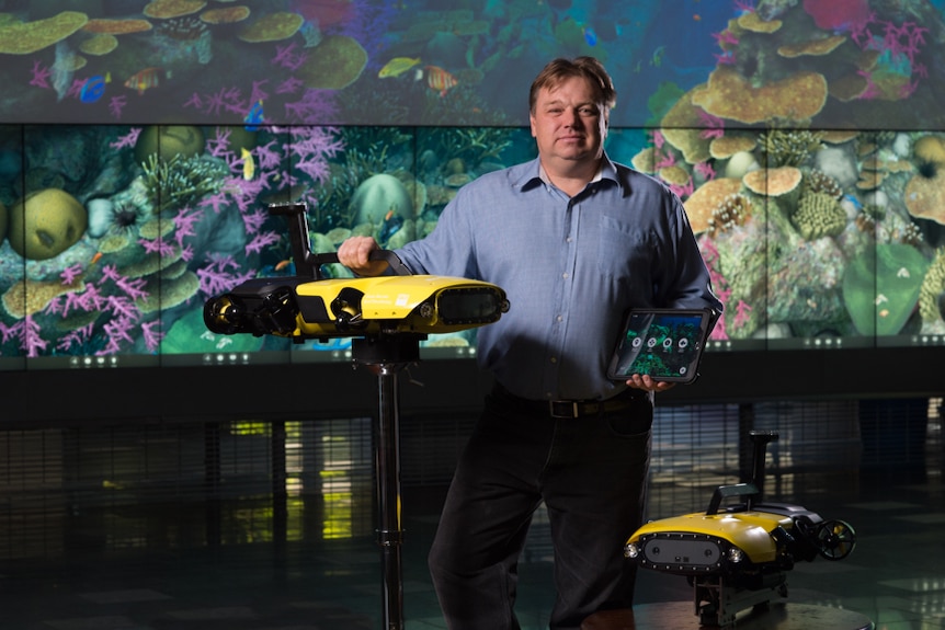 Matt Dunbabin stands next to two yellow robots in a room with a coral picture backdrop.