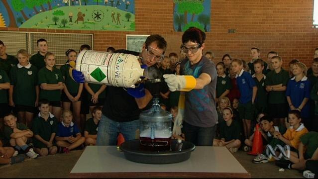 Two men wearing safety gloves and glasses pour liquid nitrogen into vessel, as school kids watch in background
