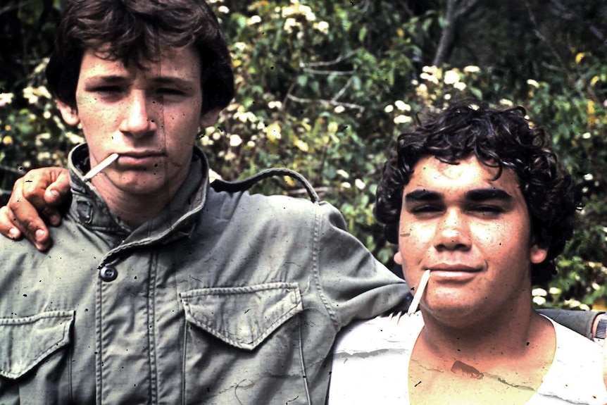 Two teenage boys stand shoulder-to-shoulder with cigarettes in their mouths.