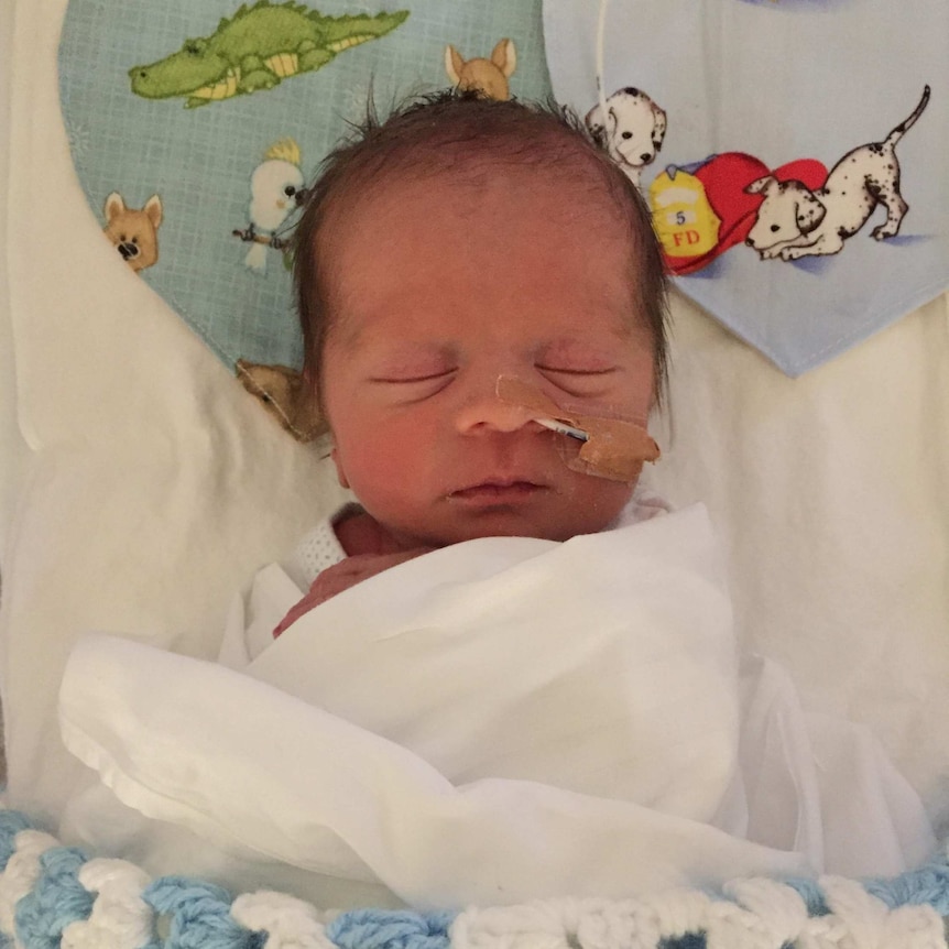 A newborn baby in a cot. He has a tube in his nose and is wrapped in a blue blanket.