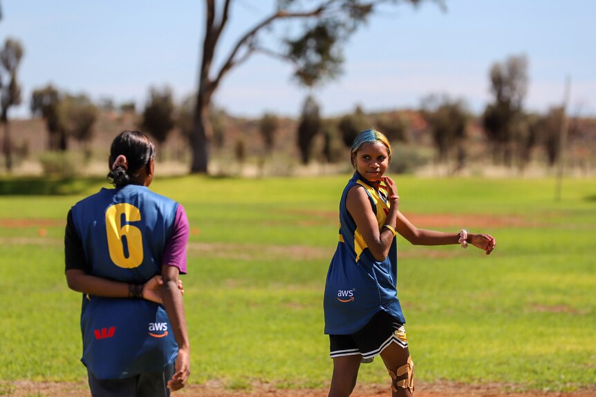 A young Aboriginal woman wearing football guernsey smiles at team mate on field