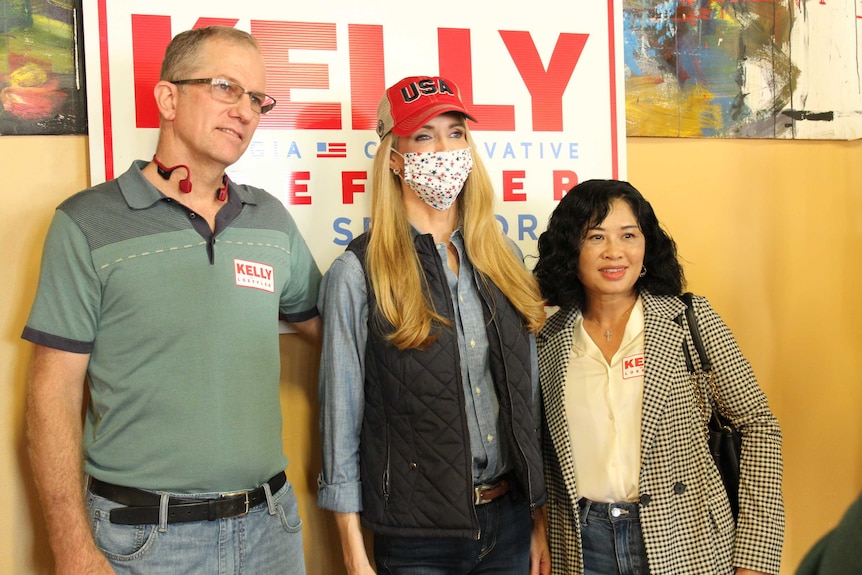 Two women and a man pose in front of a sign saying Kelly.
