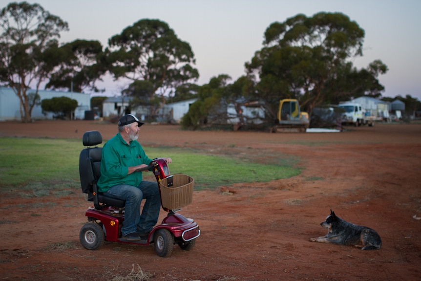 A man on a mobility scooter in the red earth driveway of a property. A dog lies on the ground in front of him.