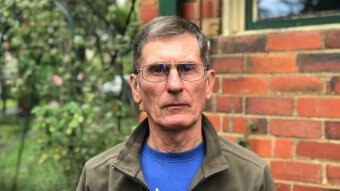 James Ashburner wears a blue t-shirt and green jacket as he stands in front of a brick home.