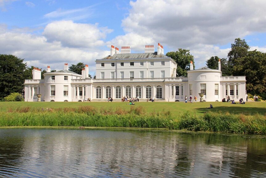 A white neoclassical building is seen from a lake which reflects the building in the water.