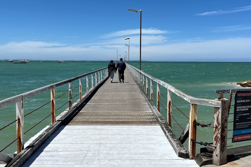 A man and a woman hold hands as they walk down a jetty.