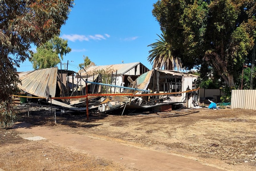 A house in ruins after fire. Police tape surrounds the wreckage and there are trees on either side.