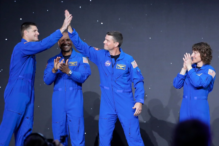 Two astronauts high five as the four stand on stage in blue suits.