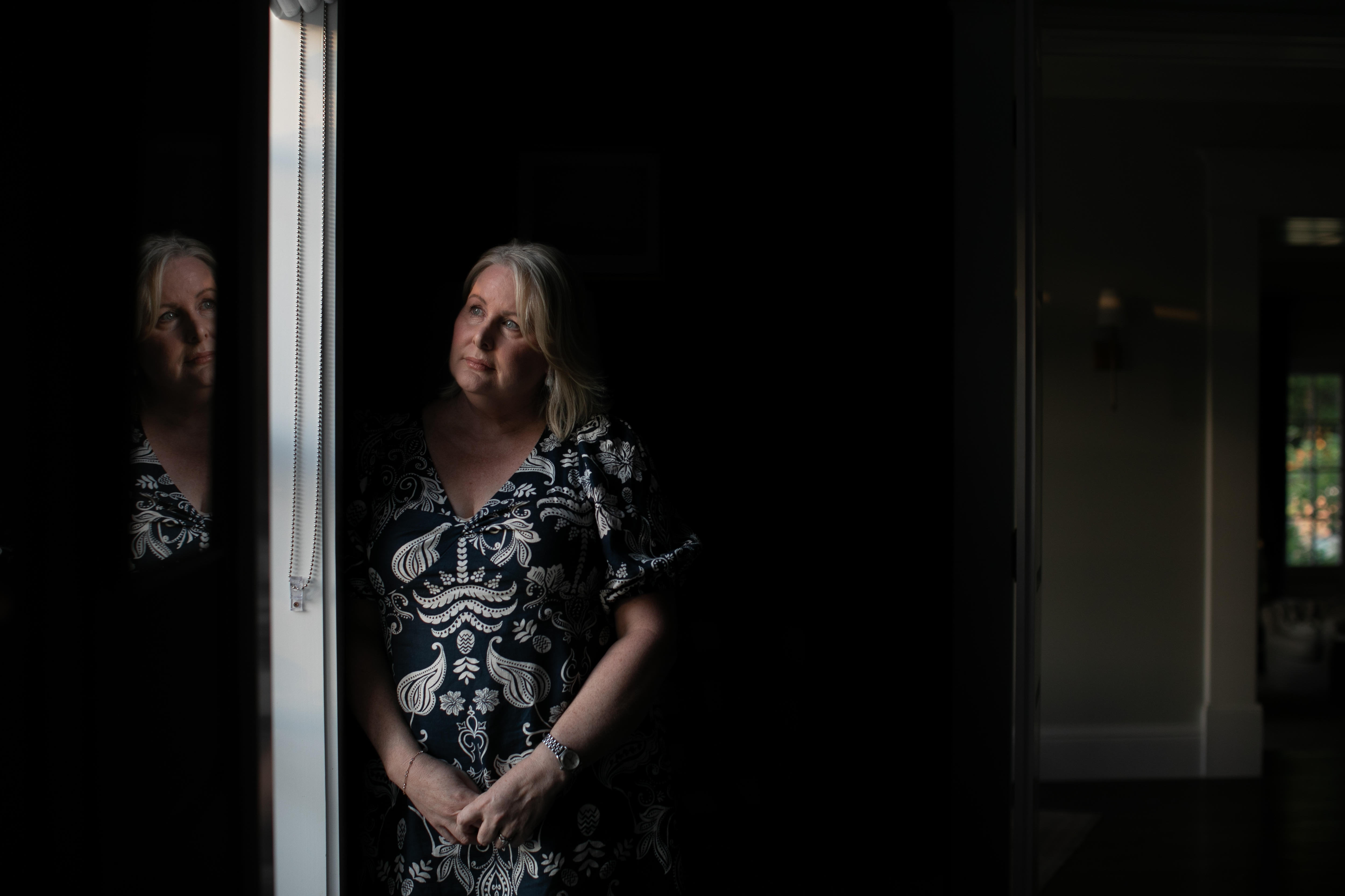 A woman wearing patterned dress stands by a window and gazes out, her reflection looking back