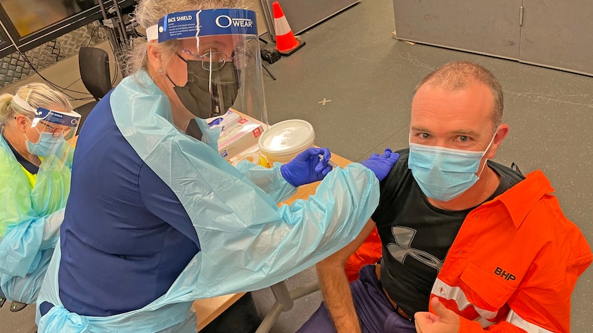 Man in fluro work gear holds thumbs up as he receives a vaccine from woman in PPE 
