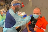 Man in fluro work gear holds thumbs up as he receives a vaccine from woman in PPE 
