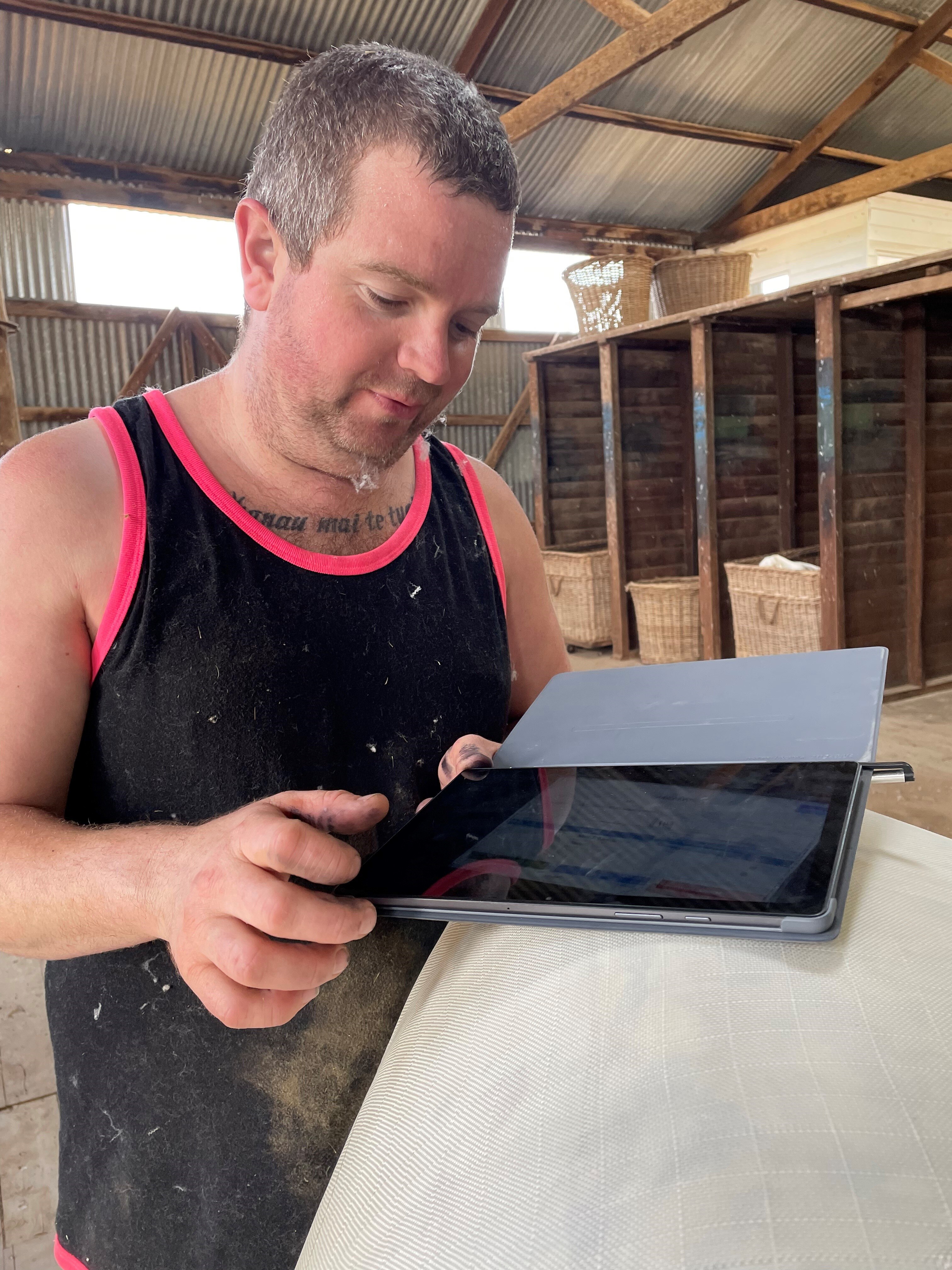A man in a singlet uses an iPad on top of a wool bale in a shearing shed 