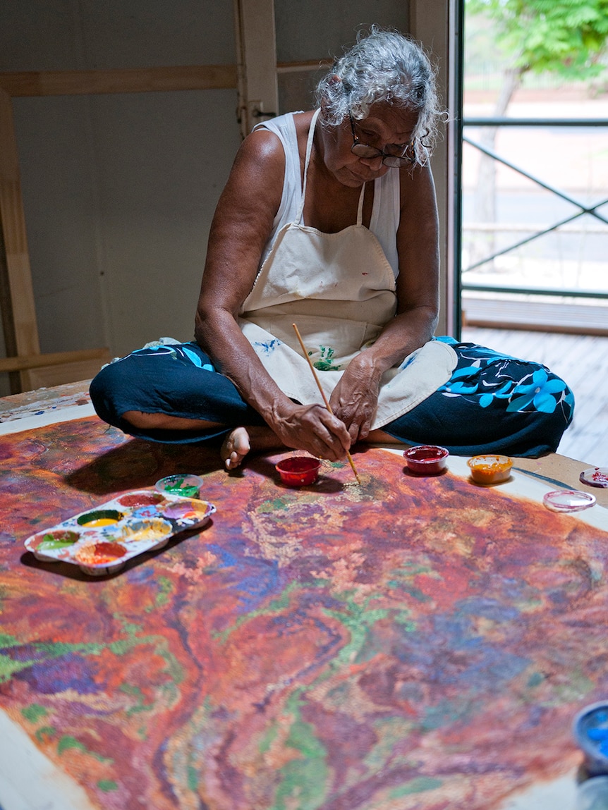 A woman with grey hair and glasses sits bends over table and paints with kebab stick on colourful dotted canvas in studio.