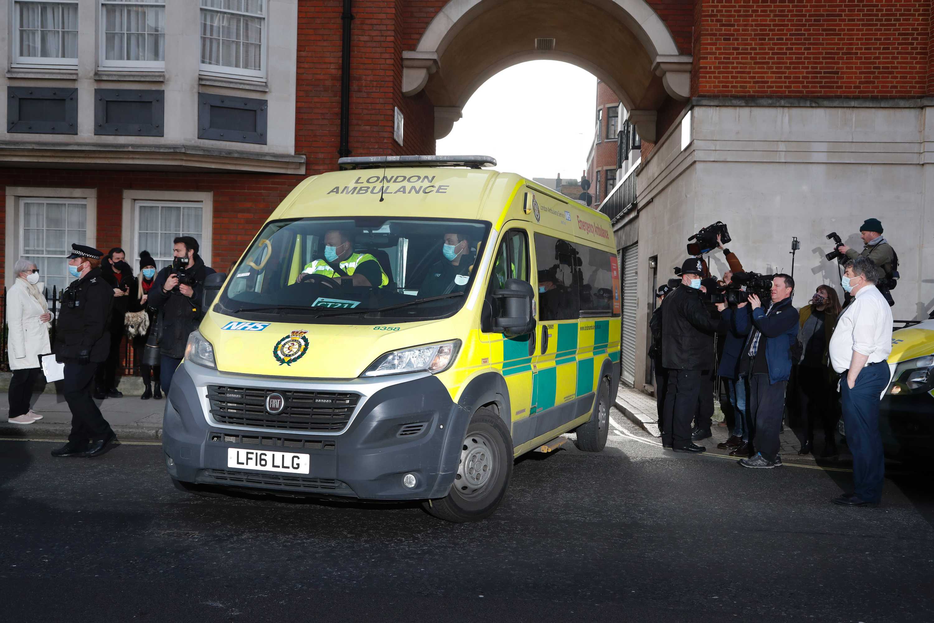 An ambulance drives out of a hospital building surrounded by media.