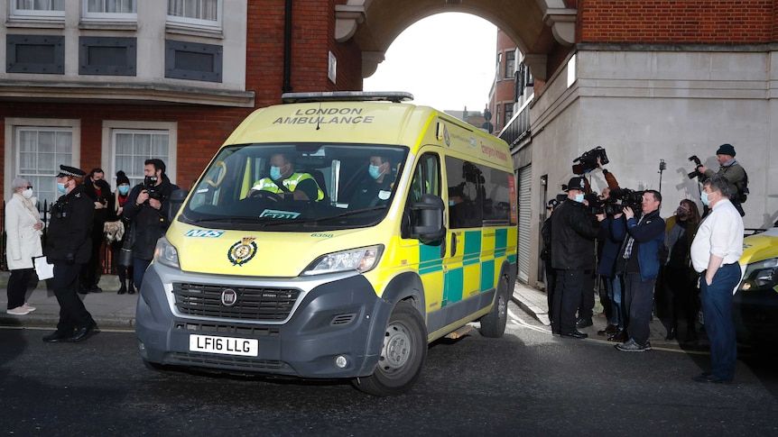 An ambulance drives out of a hospital building surrounded by media.