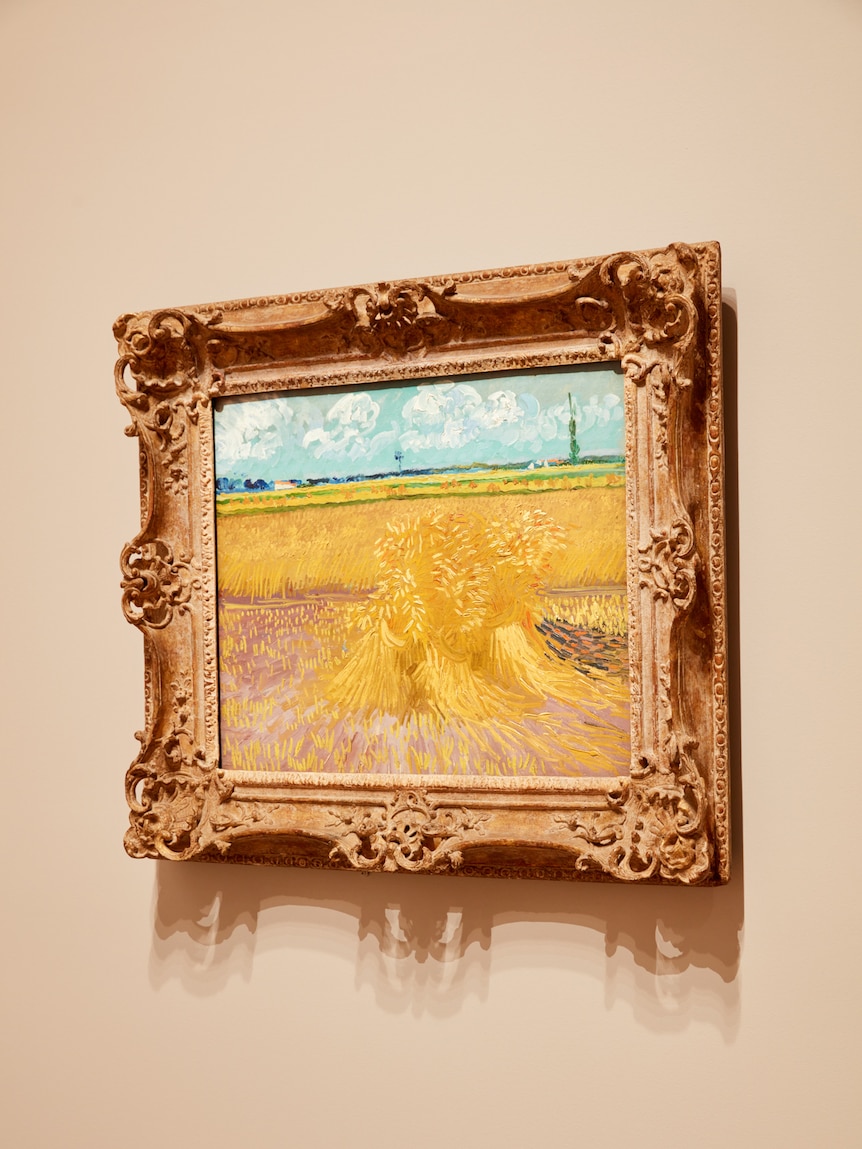 Grey wall with painting of yellow sheaf of wheat hung on it, in a gilt frame.