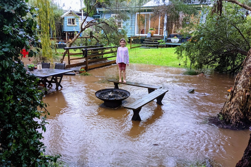 A young girl stands on an outdoor benhc in a flooded backyard with a house behind her.