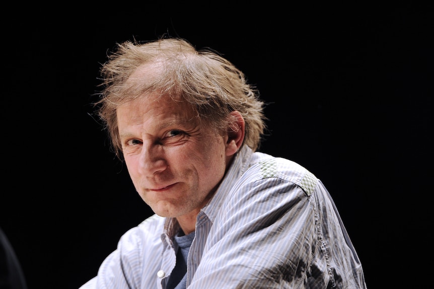 Simon McBurney, a middle-aged white man with scruffy grey hair and brown eyes, sits wearing a pale blue shirt.