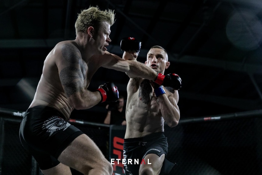 In a boxing ring, Sam Marles, in shorts, wearing boxing gloves, swings a punch past another man, also punching.