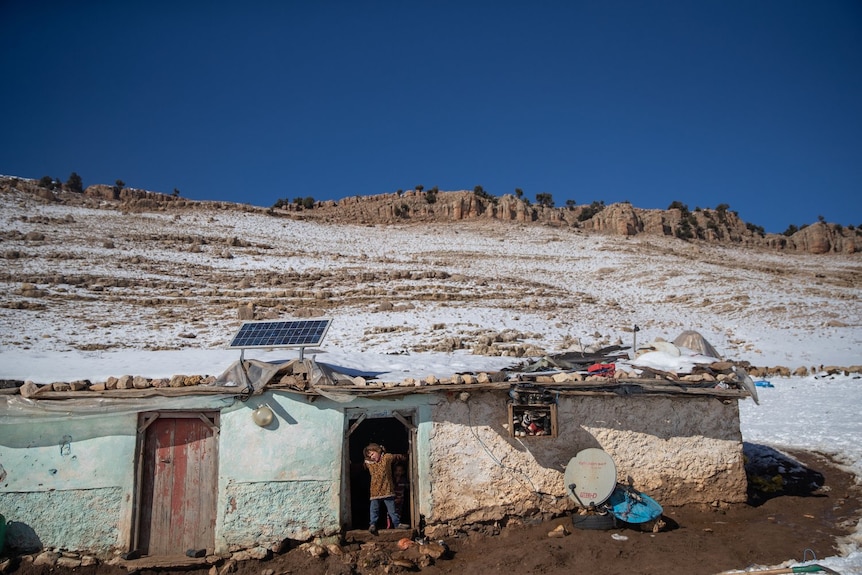 A child stands in the doorway of a simple home, with snow-covered mountains in the background.